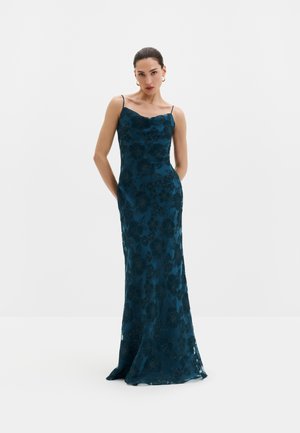 Vrouw draagt een lange donker teal bloemen slipjurk met dunne bandjes en gouden creolen, staat met haar handen achter haar rug.