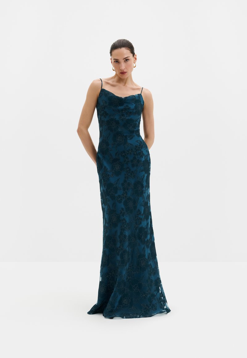 Femme portant une longue robe nuisette florale bleu sarcelle foncé avec de fines bretelles et des boucles d'oreilles créoles dorées, debout les mains derrière le dos.