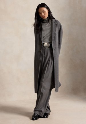 Femme portant un long manteau gris par-dessus un pull à col roulé gris et un pantalon gris à jambes larges, chaussures noires, debout la tête inclinée vers le bas.