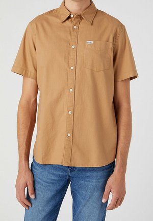 Mand iført en kortærmet beige skjorte med brystlomme og afslappede blå jeans, stående mod en ensfarvet baggrund.