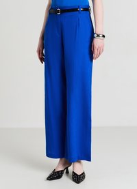 Pantalon large bleu vif en tissu lisse, avec des plis à l'avant et une ceinture noire. Associé à des chaussures à talons noires ornées de clous.