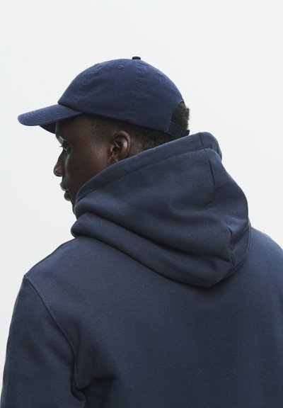 Sweat à capuche bleu marine avec une grande capuche, fabriqué à partir d'un tissu lisse, présentant une texture subtile. Porté avec une casquette assortie bleu marine.