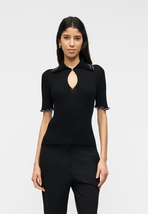 Nő hosszú sötét hajjal, fekete bordás rövid ujjú felsőt visel kulcslyuk nyakkivágással és fekete nadrágot semleges háttér előtt.