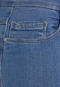 Primer plano de unos jeans de mezclilla azul que muestran un bolsillo trasero con costura naranja visible y un remache metálico en el lado.