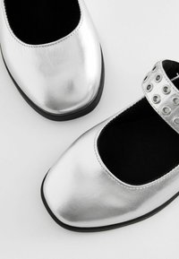 Stříbrné kožené baleríny Mary Jane s kulatou špičkou, černým vnitřním podšívkou a páskem se stříbrnými průchodkami na svršku.