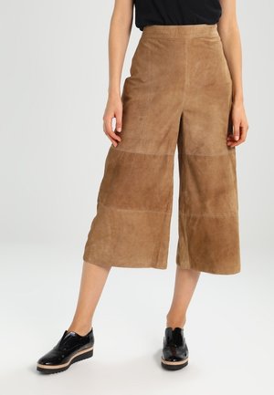 Modelo lleva pantalones anchos de ante color arena cortos y zapatos de plataforma de charol negro con suelas de madera.