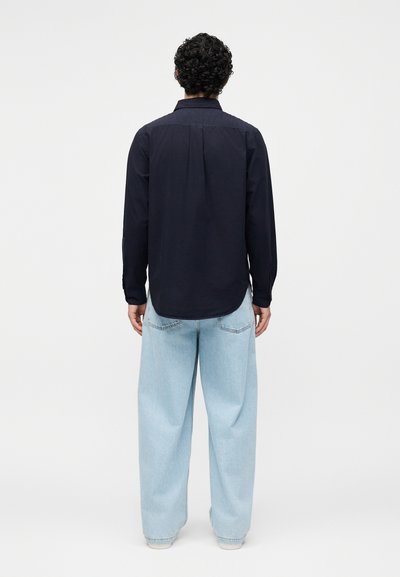 Άνδρας με σγουρά μαλλιά, που φοράει μαύρο μακρυμάνικο πουκάμισο και ανοιχτό μπλε φαρδιά τζιν, στέκεται με την πλάτη προς ένα απλό φόντο.