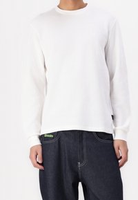 Weißes, langärmliges Thermoshirt mit gerippter Struktur, Rundhalsausschnitt und kleinem Logo auf der Brust. Kombiniert mit dunklen Jeans aus Denim mit grünem Etikett.