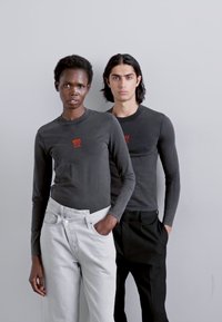 Zwei Models tragen dunkle Langarmshirts mit einem kleinen roten Logo. Das männliche Model kombiniert sein Shirt mit schwarzen Hosen, während das weibliche Model helle Jeans trägt.