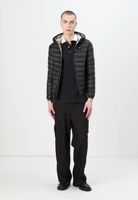 Giacca a piuma nera con cappuccio, dotata di chiusura a zip e fodera grigio chiaro. Abbinata a pantaloni cargo neri e scarpe scure.