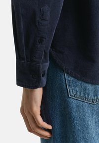 Mano sinistra rilassata lungo il fianco, indossa una camicia a maniche lunghe in velluto a coste blu scuro e jeans blu con cuciture della tasca posteriore visibili.