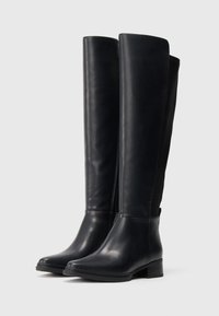 Paire de bottes en cuir noir hautes avec de petits talons blocs, fermeture éclair latérale et finition lisse sur un fond clair.