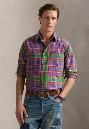 Νέος άνδρας που φοράει πουκάμισο με μανίκια γυρισμένα, μωβ και πράσινο καρό, τζιν με κομμάτια υφάσματος, καφέ ζώνη και ρολόι χειρός με δερμάτινο λουράκι.