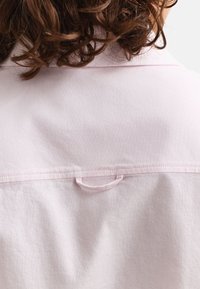 Short curly brown hair above the collar of a light pink shirt showing a fabric loop at the upper back.