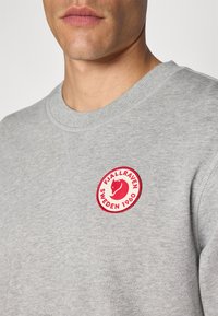 Šedá mikina vyrobená z bavlneného zmesi, s okrúhlym červeným nášivkom s logom líšky a textom "FJALLRAVEN SWEDEN 1960" na hrudi.