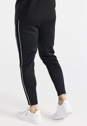 Person iført sorte, koniske bukser med hvide sidelister og hvide sneakers, der står med det ene ben let bøjet.