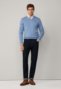 Maglione azzurro chiaro a scollo a V, jeans di denim scuro e scarpe in suede marroni. Il maglione presenta un piccolo logo e una texture liscia.