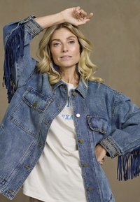 Jeansowa kurtka z niebieskim frędzlem, złotymi elementami metalowymi, dwiema przednimi kieszeniami i zapięciem na guziki. Noszona na białej koszulce z grafiką.