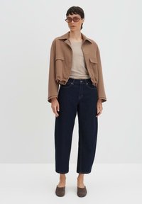 Bruine cropped bomberjack, beige top, donkerblauwe high-waisted jeans en taupe instapschoenen. Model poseert tegen een lichte achtergrond.