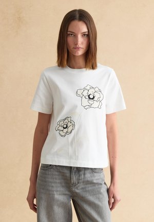 Vrouw met stijlbruin haar, gekleed in een wit T-shirt met twee zwart-wit omlijnde bloemmotieven en grijze spijkerbroek.