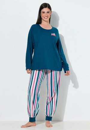 Frau steht barfuß, trägt ein türkisfarbenes Langarmshirt und gestreifte Schlafanzughosen mit rosa, türkisfarbenen und weißen vertikalen Streifen vor einem einfarbigen Hintergrund.