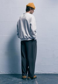 Felpa grigia dal taglio rilassato, pantaloni grigi scuri e berretto giallo senape testurizzato. Il modello si appoggia a una parete chiara.