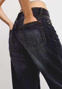 Person som bär urblekta svarta jeans med händerna i bakfickorna, kombinerat med en vit cropped-topp mot en enkel bakgrund.