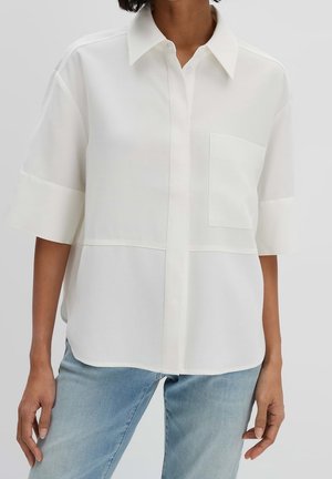 Kvinde iført løs, hvid, kortærmet skjorte med krave, knapper og brystlomme, samt lyseblå jeans, stående mod en ensfarvet baggrund.