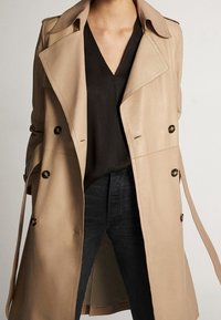 Trench-coat beige en tissu lisse, avec des boutons croisés, un large col et des sangles de taille ajustables, associé à un haut noir et un jean foncé.