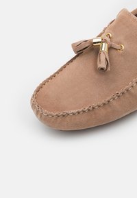 Suede moccasin shoe in light brown with a rounded toe, decorative tassels, and gold-tone hardware accenting a simple lace design.
