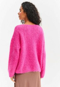 Rozmyty różowy sweter z szerokim dekoltem, opuszczonymi ramionami i długimi rękawami. Faktura materiału dodaje miękkości; linia dołu sięga talii.