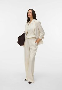 Kremowy, dopasowany żakiet i szerokie spodnie, wraz z dopasowaną koszulką polo. Strój cechuje się gładką teksturą i ciemną torebką.