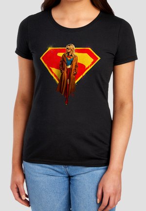 Woman wearing black t-shirt with Supergirl graphic showing her in costume and brown trench coat in front of red and yellow emblem.