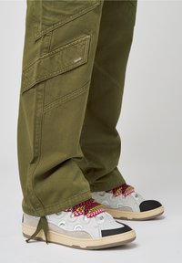 Pantalons cargo verts olive avec poches latérales et poignets, assortis à des baskets blanches avec des lacets colorés et à motifs et une coque noire.
