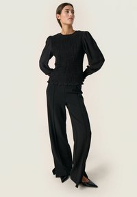 Black smocked top with long balloon sleeves, paired with high-waisted black trousers and pointed-toe black heels. Smooth texture throughout.