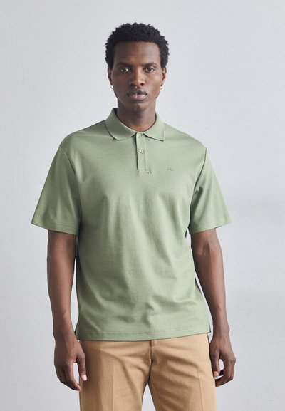 Polo verde deschis cu guler clasic, mâneci scurte și un logo subtil. Purtați cu pantaloni bej, cu o textură fină.