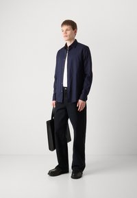 Cămașă albă cu nasturi navy, tricou alb dedesubt, blugi negri relaxați, geantă neagră și pantofi negri, stând în fața unui fundal deschis.