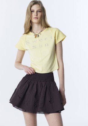 Junge Frau in einem gelben "Sweet Sun" bauchfreien Oberteil und einem dunkelbraunen Rock mit floralen Ausschnitten, die eine Charms-Halskette trägt, vor einem schlichten weißen Hintergrund.