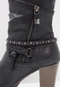 Botte en cuir noir mi-mollet avec fermeture éclair latérale, bride cloutée autour de la cheville, détail de lacet noué et talon en bois superposé.