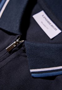 Marinblå polo t-shirt med dragkedja i kragen, med texturerad tyg och kontrasterande vita och ljusblå ränder på ärmarna. Varumärkesetikett synlig.