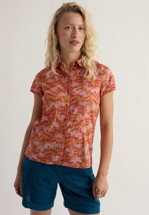 Blondynka w pomarańczowej, kwiecistej, krótkorękawowej koszuli na guziki oraz ciemnoniebieskich, podwiniętych krótkich spodenkach na jasnym tle.