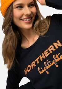 Czarna koszula z długim rękawem z pomarańczowym haftowanym napisem "NORTHERN highlights". Model nosi pomarańczową dzianą czapkę. Gładka faktura materiału.
