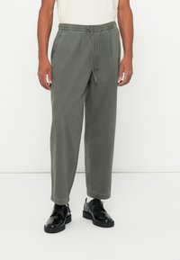 Let beanskede, løse olivengrønne bukser med elastisk talje, snøre detaljer, manchetter og en glat tekstur, parret med sorte sko.