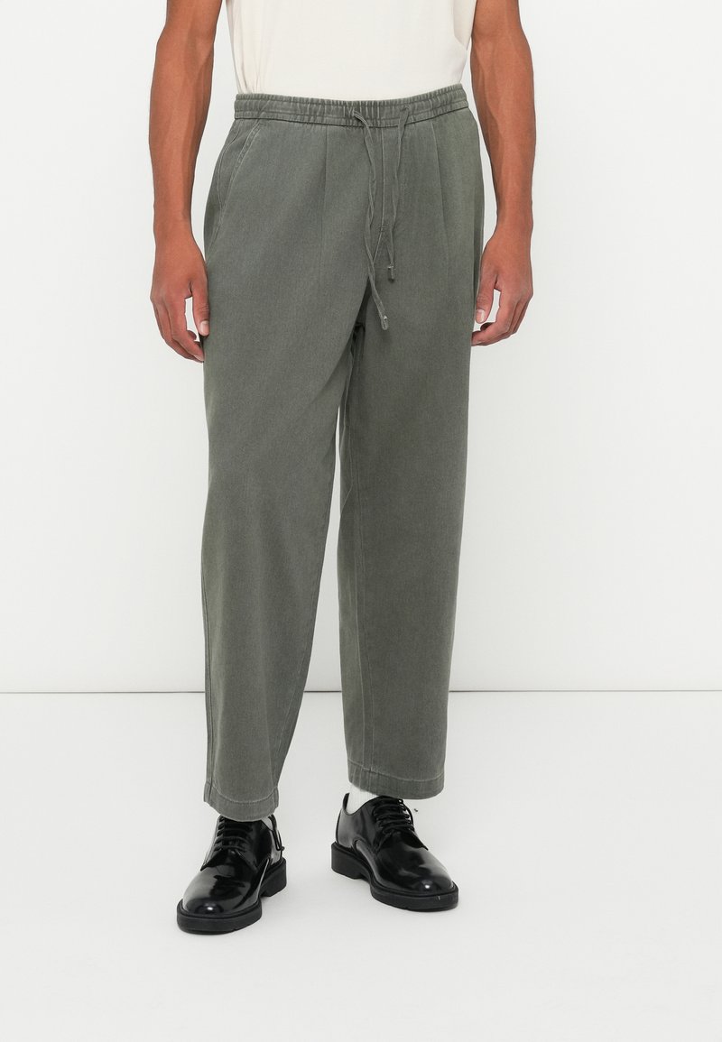 Let beanskede, løse olivengrønne bukser med elastisk talje, snøre detaljer, manchetter og en glat tekstur, parret med sorte sko.