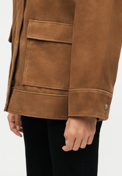 Καφέ μανίκι σακάκι από σουέτ με τσέπη με καπάκι και μανσέτα με κουμπί σε πρόσωπο που φοράει μαύρο παντελόνι σε λευκό φόντο.