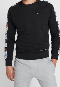 Felpa nera con toppe colorate del logo della squadra lungo le maniche; presenta un collo rotondo, polsini a costine e un'etichetta con il logo all'orlo.