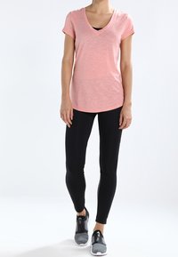 Světle růžové tričko s výstřihem do V a krátkým rukávem, spárované s černými legínami a šedými sportovními botami. Materiál se jeví jako měkký, design je neformální.