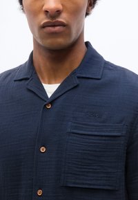 Homme portant une chemise bleu marine à boutons avec une poche poitrine et un logo Schott NYC discret brodé au-dessus de la poche.