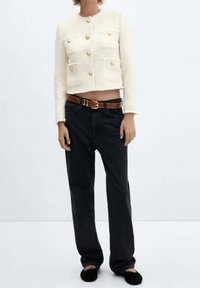 Veste courte blanche texturée avec boutons dorés et deux poches poitrine, associée à un jean noir ample et des chaussures plates noires. Ceinture marron incluse.