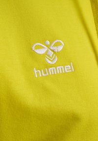 Helles gelbes Gewebe mit einem weißen gestickten Logo mit abstraktem Design und dem Text "hummel" darunter. Glatte Textur, minimalistischer Stil.